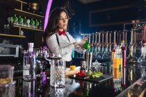 Молодая барменша в стильном наряде, сжимающая лимон, пока готовит коктейль, стоя у стойки в современном баре — стоковое фото