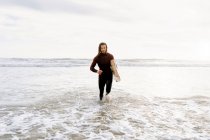 Surfer uomo vestito con muta in esecuzione con tavola da surf sulla spiaggia durante l'alba — Foto stock