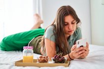 Вид сбоку молодой студентки, просматривающей социальные сети по мобильному телефону рядом со столом со свежими фруктами и соком, проводя утро дома — стоковое фото