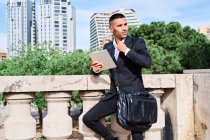 Side view of focused young Hispanic male executive manager in classy suit with bag reading information on tablet while standing near railing on urban terrace — Stock Photo