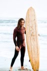 Joven surfista hombre de pelo largo vestido con traje de neopreno de pie mirando a la cámara con tabla de surf hacia el agua para coger una ola en la playa durante el amanecer - foto de stock