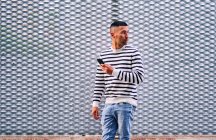 Hombre hispano adulto con ropa casual de pie cerca de la pared ornamental gris y navegando por las redes sociales en el teléfono inteligente en la calle de la ciudad - foto de stock