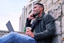 Vista lateral do homem hispânico elegante com café falando no telefone e usando netbook enquanto sentado em passos e trabalhando em projeto remoto na rua da cidade — Fotografia de Stock