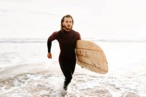 Surfista vestido de fato de mergulho correndo com prancha na praia durante o nascer do sol — Fotografia de Stock