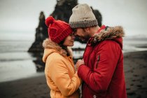 Seitenansicht junger Mann und Frau in Winterbekleidung, die an der Küste in der Nähe von großen Steinen und Wasser stehen — Stockfoto