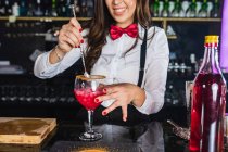 Cropped unrecognizable female barkeeper in stylish outfit stirring cocktail in a glass with long spoon standing at counter in modern bar — Stock Photo