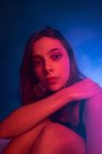 Tranquilo jovem modelo feminino no vestido sentado no chão e inclinado na mão enquanto olha para a câmera no estúdio escuro com luzes coloridas — Fotografia de Stock