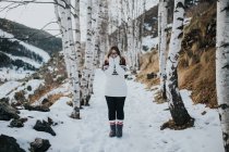 Giovane turista felice in occhiali e maglione guardando la fotocamera e in piedi tra i boschi nella neve vicino alla montagna — Foto stock