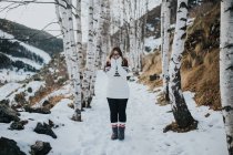 Joven turista feliz en gafas y suéter mirando a la cámara y de pie entre los bosques en la nieve cerca de la montaña - foto de stock