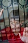 Через склянку нещасного хлопчика з синцями на обличчі, який дивиться далеко, стоячи біля вікна вдома як концепція домашнього насильства та жорстокого поводження з дітьми — стокове фото