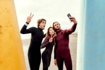 Gruppo di amici surfisti felici vestiti con mute in piedi vicino alle tavole da surf mentre si scattano selfie con smartphone sulla spiaggia durante l'allenamento — Foto stock
