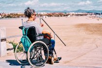 Vista lateral de la hembra discapacitada en máscara protectora sentada en silla de ruedas con mochila y disfrutando de un soleado día de verano en la playa de arena - foto de stock