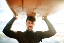 Retrato do jovem surfista vestido de fato de mergulho em pé olhando para a câmera na praia com a prancha de surf acima da cabeça durante o nascer do sol — Fotografia de Stock