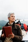 Elderly female traveler with short gray hair leaning on car and browsing tablet during road trip in countryside and looking away — Stock Photo
