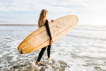 Vista laterale della donna surfista vestita in muta che cammina mentre porta la tavola da surf sopra la testa sulla spiaggia durante l'alba sullo sfondo — Foto stock