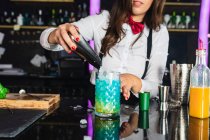 Cropped unrecognizable female barkeeper in stylish outfit adding ice cubes into a glass while preparing blue blue cocktail standing at counter in modern bar — Stock Photo