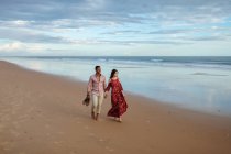 Веселая многорасовая пара, держащаяся за руки и идущая вдоль мокрого берега, любуясь летом морем на закате — стоковое фото