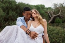 Multiracial couple of newlyweds sitting on stump of tree in woods and hugging on wedding day — Stock Photo