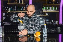 Чоловічий бармен додає апельсинову шкірку в склянку, готуючи коктейль, що стоїть за стійкою в сучасному барі — стокове фото