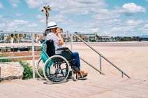 Вид збоку на невпізнавану жінку з сірим волоссям у інвалідному візку, яка фотографує фотоапарат, проводячи літній день на пляжі — стокове фото