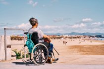 Vista lateral de la hembra discapacitada en máscara protectora sentada en silla de ruedas con mochila y disfrutando de un soleado día de verano en la playa de arena - foto de stock