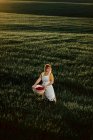 Jeune femme en robe vintage portant panier en osier tout en marchant dans un champ herbeux vert au coucher du soleil dans la campagne estivale — Photo de stock