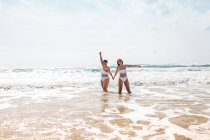 Fröhliche Freundinnen halten sich in Badeanzügen im schäumenden Meer am Sandstrand unter blauem wolkenverhangenem Himmel an sonnigen Tagen an den Händen — Stockfoto