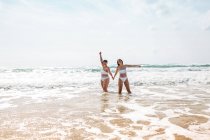 Alegre amigos femeninos tomados de la mano en trajes de baño en el océano espumoso cerca de la playa de arena bajo el cielo azul nublado en el día soleado - foto de stock