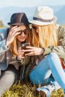 Jovens amigas em chapéus e roupas elegantes gelando no gramado verde e compartilhando telefone no campo das montanhas — Fotografia de Stock