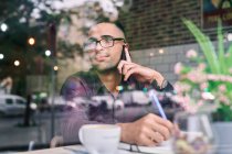 Empresario hispano inteligente en gafas tomando notas en bloc de notas y contestando llamadas telefónicas mientras está sentado detrás de la ventana en la cafetería - foto de stock