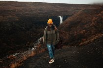 Junger Tourist mit Brille und Hut mit Piercing blickt auf Hügel in der Nähe von Wasserfall und Gebirgsfluss — Stockfoto
