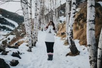 Молодой счастливый турист в очках и свитере смотрит в камеру и стоит между лесами в снегу рядом с горой — стоковое фото