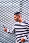 Hispanischer Mann in stylischem Outfit schaut weg und benutzt Handy, während er sich auf der Stadtstraße an die Wand lehnt — Stockfoto