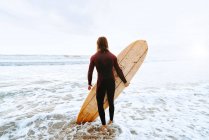 Vue de dos de surfeur méconnaissable homme vêtu d'une combinaison de plongée regardant loin avec planche de surf vers l'eau pour attraper une vague sur la plage pendant le lever du soleil — Photo de stock
