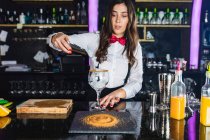 Barkeeperin in stylischem Outfit fügt Eiswürfel in ein Glas hinzu, während sie in einer modernen Bar am Tresen einen Cocktail zubereitet — Stockfoto