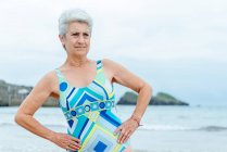 Позитивна старша жінка в стильному купальнику робить бічні вправи на вигин, практикуючи здоровий спосіб життя і працюючи на пляжі проти махаючого океану — стокове фото
