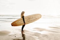 Вид збоку жінка-серфер, одягнена в гідрокостюм, стоїть озираючись з дошкою для серфінгу на пляжі під час сходу сонця на задньому плані — стокове фото