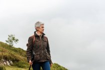 Пожилая женщина с рюкзаком, держащая трость и стоя на травянистом склоне к вершине горы во время путешествия на природе — стоковое фото