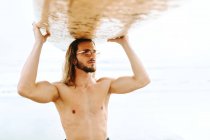 Giovane surfista uomo con i capelli lunghi vestito con muta e occhiali da sole alla moda in piedi portando la tavola da surf sulla testa guardando lontano sulla spiaggia — Foto stock