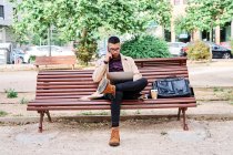 Selbstbewusster junger hispanischer Mann in stylischem lässigem Outfit und Brille, der Telefonanrufe entgegennimmt, während er auf einer Bank sitzt und mit Laptop auf der Straße der Stadt arbeitet — Stockfoto