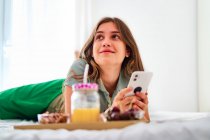 Young female student browsing social networks on mobile phone near table with fresh fruits and juice while spending morning at home — Stock Photo