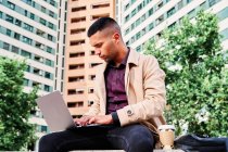 Low angle of focused young Hispanic man in stylish casual clothes working remotely on freelance project on laptop while sitting with cup of coffee near modern urban buildings — Stock Photo