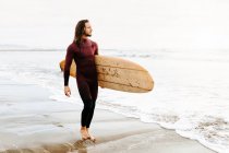 Surfer uomo vestito in muta a piedi guardando lontano con la tavola da surf verso l'acqua per catturare un'onda sulla spiaggia durante l'alba — Foto stock
