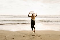 Vista posteriore di surfista donna irriconoscibile vestita in muta che cammina mentre tiene la tavola da surf sulla testa sulla spiaggia durante l'alba sullo sfondo — Foto stock