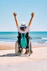 Visão traseira de viajante feminino irreconhecível em cadeira de rodas com mochila levantando braços enquanto desfruta de viagem de verão na praia perto do mar azul — Fotografia de Stock