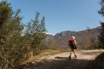 Vista trasera del explorador con mochila caminando por un camino arenoso que conduce a las tierras altas en un día soleado - foto de stock