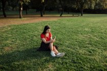 Femme en vêtements d'été assis sur la prairie verte dans le parc et surfer sur Internet sur téléphone mobile tout en divertissant le week-end dans la soirée — Photo de stock