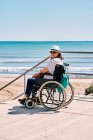 Вид збоку жінка мандрівник у інвалідному візку з рюкзаком насолоджується літньою подорожжю на пляжі біля блакитного моря, дивлячись на камеру — стокове фото