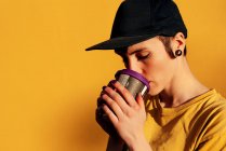 Jovem fêmea informal na moda tampão bebendo takeaway bebida quente com olhos fechados contra fundo amarelo — Fotografia de Stock