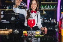 Focused female barkeeper in stylish outfit adding liquid from bottle into glass while preparing cocktail standing at counter in modern bar — Stock Photo